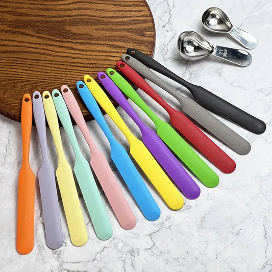 Colorful silicone spatulas on a marble surface with wooden cutting board and metal measuring spoons.