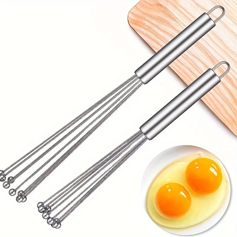 Two metal whisks on a white background with a bowl of eggs.